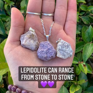 Op de afbeelding: Drie ruwe lepidolietkristal hangers aan zilveren kettingen. De kristallen zijn in verschillende tinten paars en wit. De tekst "Lepidolite can range from stone to stone" staat onder de kristallen. Twee paarse harten bevinden zich onder de tekst.