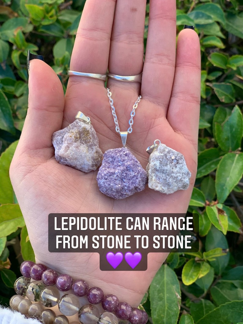 Op de afbeelding: Drie ruwe lepidolietkristal hangers aan zilveren kettingen. De kristallen zijn in verschillende tinten paars en wit. De tekst "Lepidolite can range from stone to stone" staat onder de kristallen. Twee paarse harten bevinden zich onder de tekst.