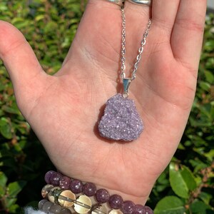 Op de afbeelding: Een zilveren ketting met een hanger van paarse lepidolietkristal. De hanger is een natuurlijke, ruw geslepen steen met een glinsterende, iriserende afwerking.