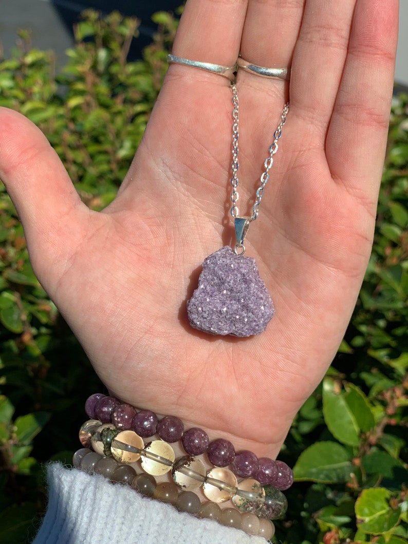 Op de afbeelding: Een zilveren ketting met een hanger van paarse lepidolietkristal. De hanger is een natuurlijke, ruw geslepen steen met een glinsterende, iriserende afwerking.