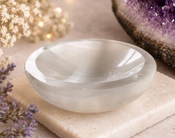Könnte beinhalten: Eine kleine, weiße Selenit-Schale mit glatter, polierter Oberfläche. Die ovale Schale steht auf einem quadratischen, hellfarbigen Stein. Der Hintergrund umfasst eine violette Amethyst-Geode und zarte weiße Blüten.