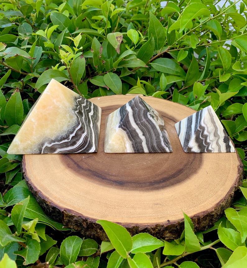 May include: Three decorative onyx pyramids with a layered pattern of cream, black, and grey. The pyramids are displayed on a round wooden slab, surrounded by green foliage. The onyx pyramids vary in size.