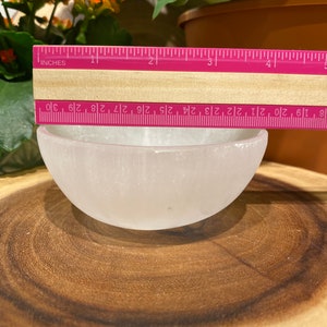 May include: A small, white selenite bowl, approximately 7.6 cm in diameter, is on a wooden surface. A pink ruler with inch markings is placed across the bowl's opening. The bowl is smooth and translucent, with a rounded shape.
