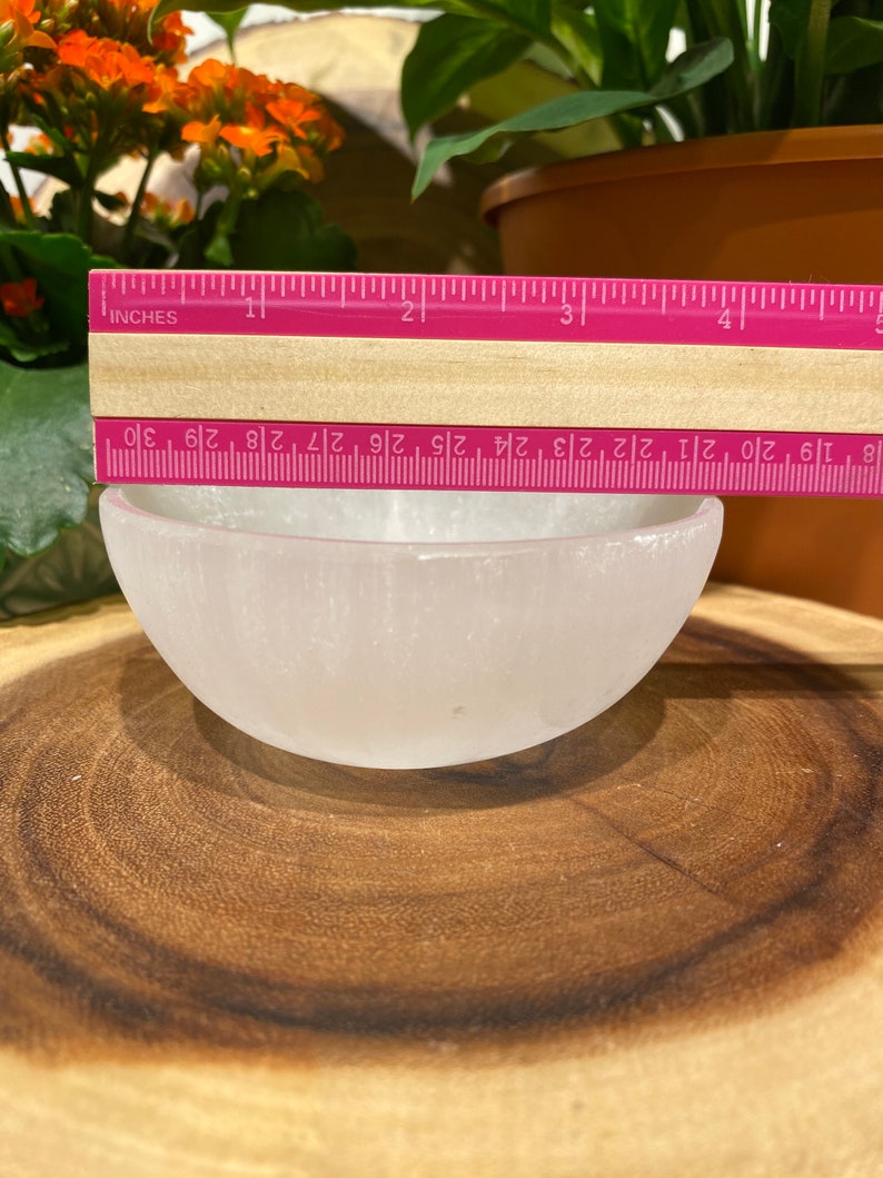 May include: A small, white selenite bowl, approximately 7.6 cm in diameter, is on a wooden surface. A pink ruler with inch markings is placed across the bowl's opening. The bowl is smooth and translucent, with a rounded shape.