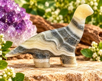 May include: A carved stone dinosaur figurine featuring horizontal bands of cream, gray, and black mineral layers. The smooth, polished surface displays natural wavy patterns throughout the body, neck, and tail, with a solid cream-colored head.