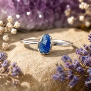 May include: A silver ring featuring a raw, oval-shaped blue gemstone. The ring is set on a light-colored surface, surrounded by purple and white flowers. The gemstone has a textured appearance.