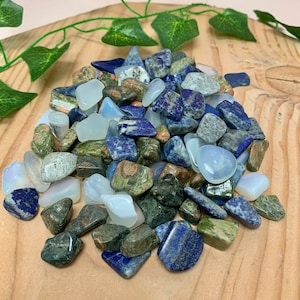 May include: A pile of polished gemstones in various colors, including deep blue lapis lazuli, milky white opal, and speckled green stones. The stones are arranged on a light brown wooden surface, with green vine accents.