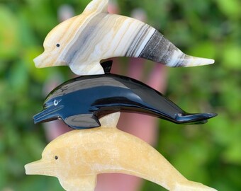 May include: Three carved dolphin figurines stacked on top of each other. The top dolphin is made of white and brown onyx, the middle dolphin is black, and the bottom dolphin is a light yellow color.