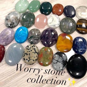 May include: A collection of polished worry stones in various colors and patterns, including green, blue, pink, and speckled designs. The stones are oval-shaped and arranged on a light-colored surface. Text at the bottom reads "Worry stone collection."
