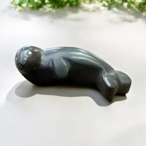 May include: A hand-carved gray stone sculpture of a seal lying on its belly. The sculpture is detailed with realistic features, including the seal's flippers and whiskers.