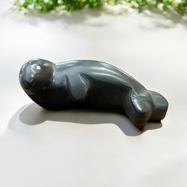 May include: A hand-carved gray stone sculpture of a seal lying on its belly. The sculpture is detailed with realistic features, including the seal's flippers and whiskers.