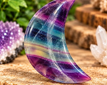 May include: A crescent moon-shaped carving made of polished fluorite stone. The mineral displays distinct horizontal bands of deep purple, vibrant teal, and pale yellow colors with natural internal fractures and a smooth, glossy surface finish.