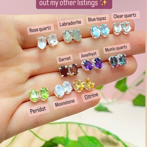 May include: A close-up of a hand holding a variety of gemstone earrings. The earrings are oval-shaped and set in silver. The stones include rose quartz, labradorite, blue topaz, clear quartz, garnet, amethyst, mystic quartz, peridot, moonstone, and citrine.