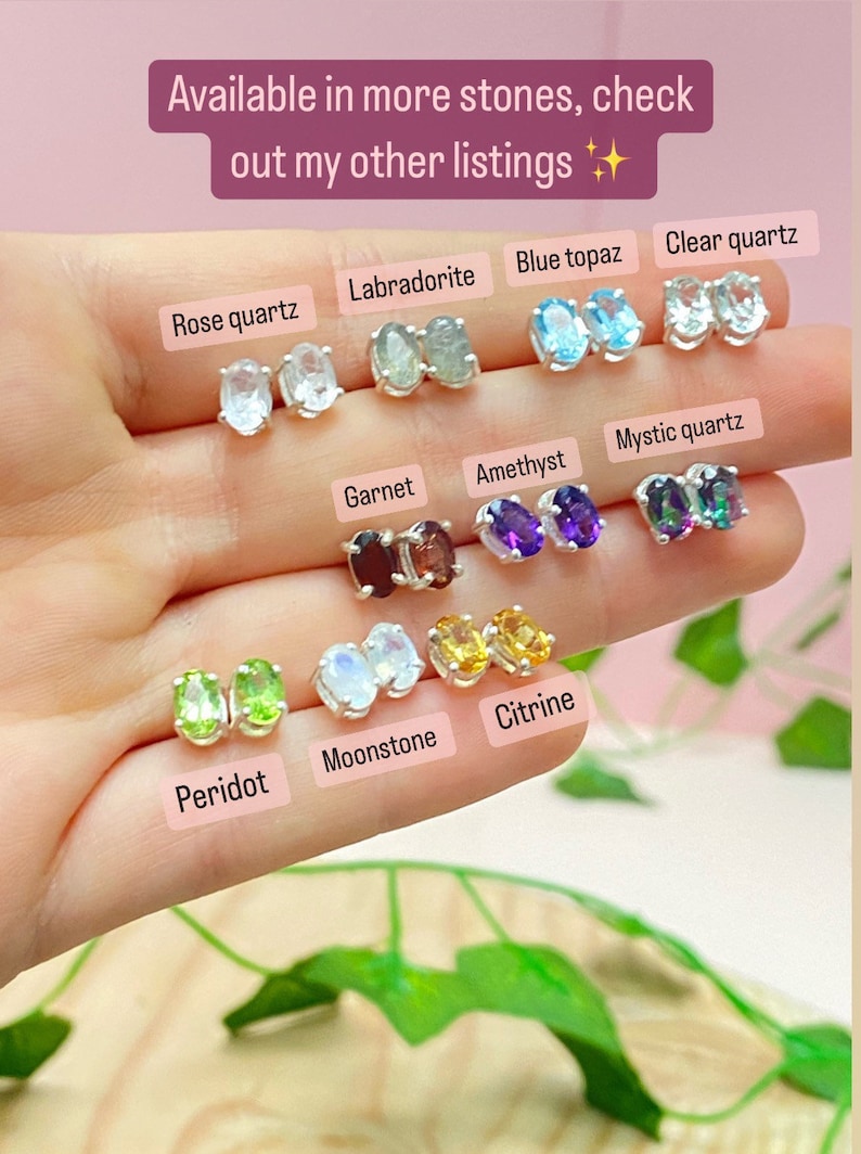 May include: A close-up of a hand holding a variety of gemstone earrings. The earrings are oval-shaped and set in silver. The stones include rose quartz, labradorite, blue topaz, clear quartz, garnet, amethyst, mystic quartz, peridot, moonstone, and citrine.