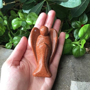 May include: A carved sandstone angel figurine in a reddish-brown colour. The angel is standing with its wings outstretched and its hands clasped in prayer.