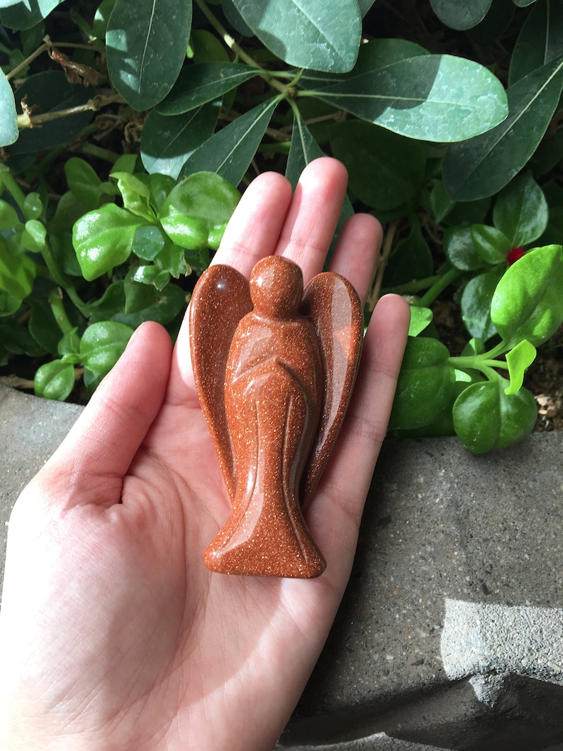 May include: A carved sandstone angel figurine in a reddish-brown colour. The angel is standing with its wings outstretched and its hands clasped in prayer.
