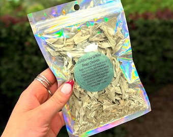 May include: A clear, iridescent bag filled with dried California White Sage leaves. The bag has a circular sticker with text describing the sage's origin and uses. The sage is a light green color.