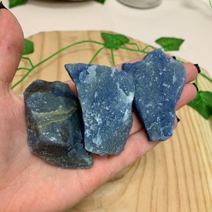 May include: Three blue and gray rough-cut pieces of blue quartz crystal. The crystals are on a hand.