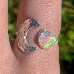 May include: A silver crescent moon ring with a pink opal gemstone. The ring is on a finger.