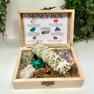 Peut inclure: Bo&icirc;te en bois avec l'inscription "Money Box" contenant une vari&eacute;t&eacute; de cristaux et un b&acirc;tonnet de sauge. La bo&icirc;te comprend des cristaux de quartz clair, d'am&eacute;thyste, de cornaline, de pyrite, d'&eacute;meraude, d'am&eacute;thyste et d'&oelig;il de tigre. La bo&icirc;te comprend &eacute;galement un b&acirc;tonnet de sauge.