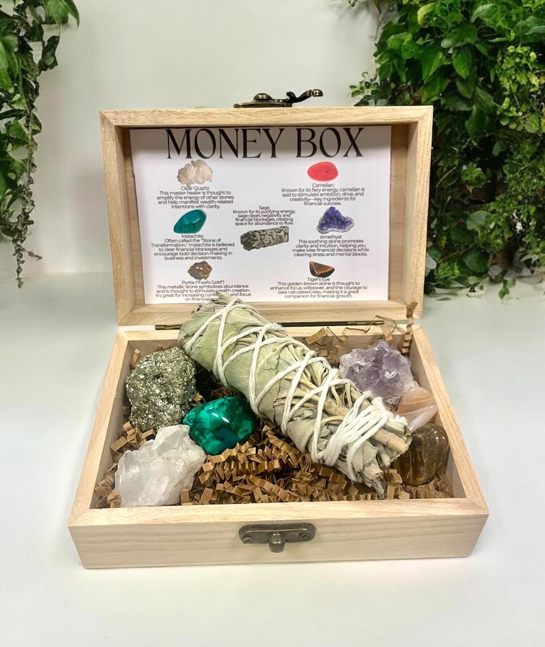 Peut inclure: Bo&icirc;te en bois avec l'inscription "Money Box" contenant une vari&eacute;t&eacute; de cristaux et un b&acirc;tonnet de sauge. La bo&icirc;te comprend des cristaux de quartz clair, d'am&eacute;thyste, de cornaline, de pyrite, d'&eacute;meraude, d'am&eacute;thyste et d'&oelig;il de tigre. La bo&icirc;te comprend &eacute;galement un b&acirc;tonnet de sauge.