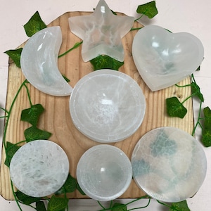 May include: A selection of white selenite crystal bowls and shapes, including a crescent moon, star, and heart. The items are displayed on a wooden board with green vine decorations. The bowls vary in size and shape, with a smooth, translucent appearance.