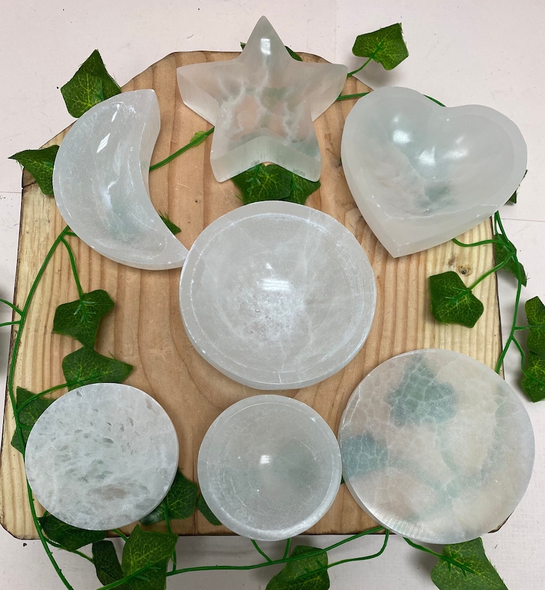 May include: A selection of white selenite crystal bowls and shapes, including a crescent moon, star, and heart. The items are displayed on a wooden board with green vine decorations. The bowls vary in size and shape, with a smooth, translucent appearance.