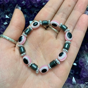 May include: A pink and black eye charm bracelet with silver beads. The bracelet is made with alternating black and silver beads, with pink and black eye charms in between.