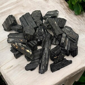 May include: A pile of black tourmaline crystals, also known as schorl, on a light wood surface. The crystals are rough and have a natural, unpolished look.