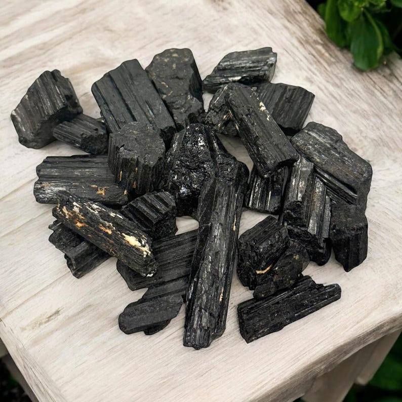 May include: A pile of black tourmaline crystals, also known as schorl, on a light wood surface. The crystals are rough and have a natural, unpolished look.