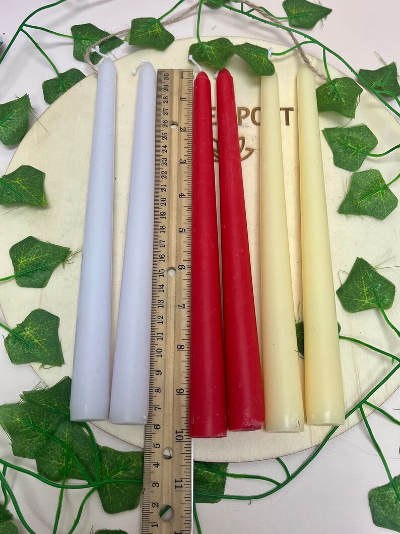 May include: Four taper candles, two white, one red, and one cream, arranged on a wooden surface with a ruler measuring 10 inches. The candles are approximately 10 inches long.