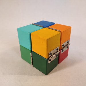 May include: A six-sided wooden cube with each side a different colour: teal, yellow, green, red, orange, and blue. Each side has a silver hinge connecting it to the adjacent side.