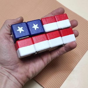 Può includere: Un braccialetto di legno con un design che ricorda la bandiera americana. Il braccialetto è composto da piccoli blocchi rettangolari dipinti di rosso, bianco e blu, con stelle bianche sui blocchi blu. Il braccialetto è tenuto in una mano.