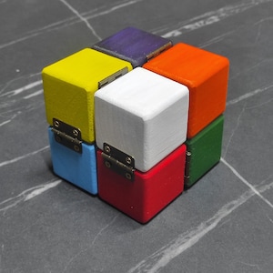 Puede incluir: Un rompecabezas de cubo de madera de colores con componentes en forma de caja con bisagras. El cubo presenta seis lados, cada uno pintado de un color diferente: amarillo, azul, rojo, blanco, naranja, verde y morado. El cubo descansa sobre una superficie gris.