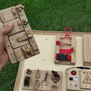 Puede incluir: Tablero de actividades de madera con varios pestillos, cerraduras e interruptores. Cuenta con una puerta de madera con bisagras y cierres metálicos, una hebilla roja y un panel con elementos de temática eléctrica. Diseñado para el juego educativo.