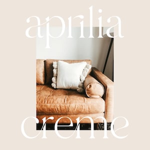 May include: A brown leather sofa with a white pillow with a fringe trim. The sofa is in a living room with a white wall.
