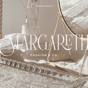 May include: A white text logo on a white background reads "MARGARETH FASHION & CO.". The logo is displayed on a glass tray with a decorative pattern. A gold metal stand with a round mirror is in the background.