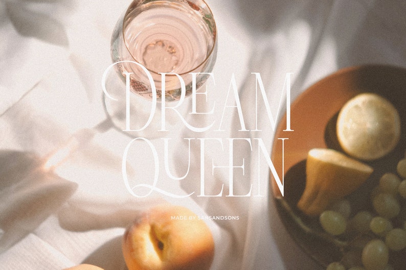 May include: A white background with a glass of pink liquid, a peach, a lemon, and a plate of grapes. The text "DREAM QUEEN" is written in white on the background. The text "MADE BY SANDSONS" is written in white below the text "DREAM QUEEN".