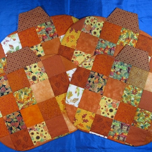 May include: Set of four orange, brown, and yellow patchwork fabric pumpkin-shaped placemats. Each placemat has a different pattern of fall leaves, sunflowers, and other autumnal designs.