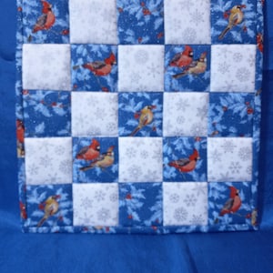 May include: Square quilted potholder with a checkerboard pattern. Alternating squares of white fabric with snowflakes and blue fabric with cardinals and holly. The edges are bound with a matching blue fabric.