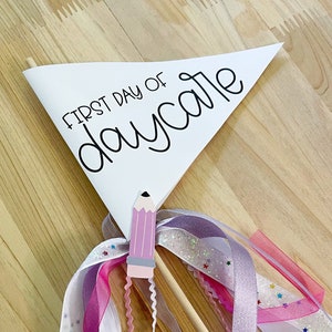 Op de afbeelding: Een witte driehoekige vlag met de tekst "First Day of Daycare" in zwarte cursieve letters. Een roze potlood met een houten stok en kleurrijke linten zijn aan de vlag bevestigd.