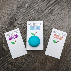 May include: Three white cards with the text "You're the Balm" in colorful letters. The middle card has a blue lip balm on top of it.