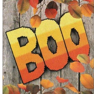 May include: A close-up of a handmade "BOO" sign made of plastic canvas with a gradient of yellow, orange, and red. The sign is surrounded by autumn leaves.