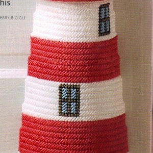 May include: A red and white striped lighthouse made of yarn with a brown roof and blue windows. The lighthouse is sitting on a gray base with two small rocks.