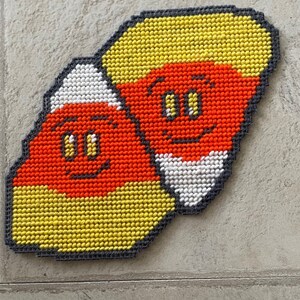 May include: Two candy corn shaped felt ornaments with smiling faces. The ornaments are orange, yellow, and white.