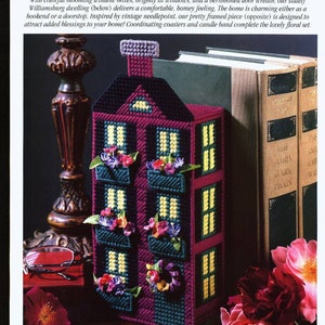 Könnte beinhalten: Ein lila und grüner Plastik-Canvas-Gehäuse für eine Tissuebox in Form eines Hauses mit einem weißen Schornstein und gelben Fenstern. Das Haus ist mit bunten Blumen und Grünpflanzen verziert. Das Tissuebox-Gehäuse steht auf zwei Büchern.