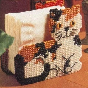 May include: A handmade tissue box cover shaped like a calico cat. The cat is made of plastic canvas with orange, black, and white yarn. The tissue box is filled with white tissues.