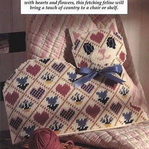 以下が含まれることがあります： クリーム色の背景に赤いハート、青い花、青いリボンが飾られた、手作りのキャリコ猫型のクッション。濃い赤色の毛糸玉が近くにあります。テキストは「Our calico kitty is a real charmer!」と書かれています。