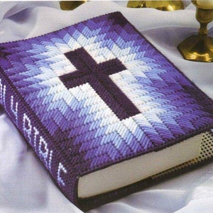 May include: A handcrafted book cover featuring a cross motif. The cover is made from yarn in shades of purple, blue, and white. The spine displays the words "HOLY BIBLE". The cross is centrally positioned and is a dark purple colour.