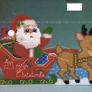 May include: A red and green plastic canvas Christmas craft featuring Santa Claus in a sleigh pulled by a reindeer. The sleigh has a white banner that reads "Merry Christmas".